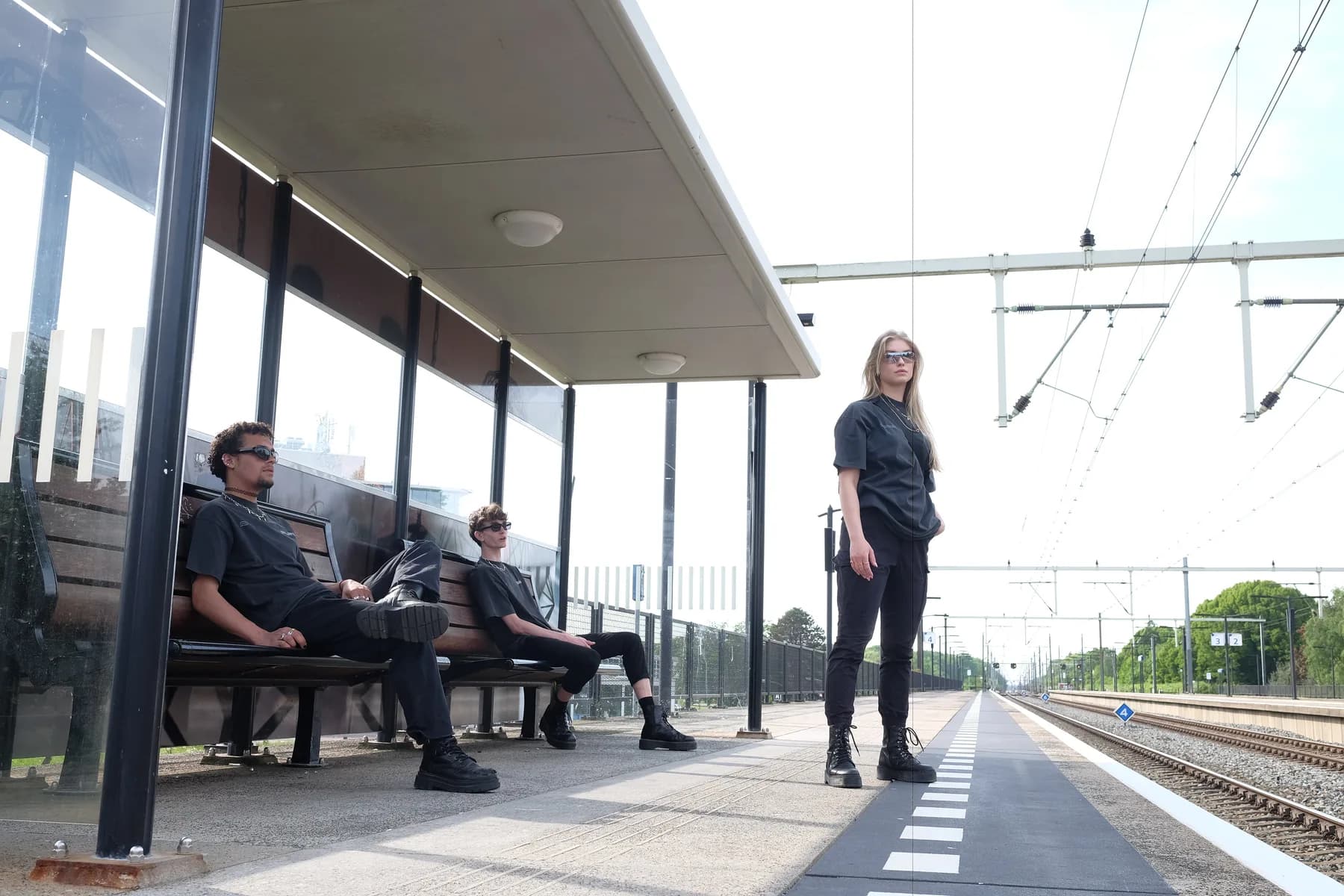 Three people in dark streetwear on a train platform under overhead wires